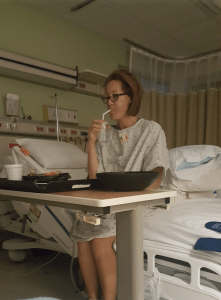A woman in a hospital gown sits on a hospital bed, drinking from a cup with a straw. A food tray is on the table in front of her. The room has medical equipment and beige curtains in the background.