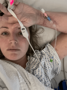 A woman in a hospital gown lies in bed with a solemn expression. An IV is inserted in her bruised arm, and a pulse oximeter glows red on her finger. Medical equipment is visible on her hand and arm.