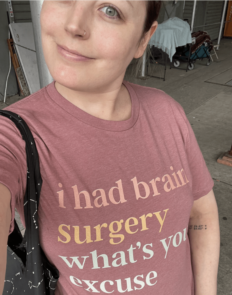 A person with light skin and a nose ring smiles while wearing a pink T-shirt that says, i had brain surgery whats your excuse. They have a black bag over one shoulder and are standing outdoors.