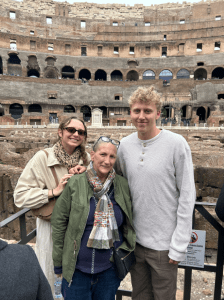 Three people posing and smiling in front of the ancient stone structure of the Colosseum in Rome, with its layered arches and weathered walls visible in the background.