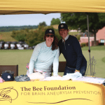 Two smiling people stand under a yellow tent at an outdoor event, behind a table with hats and shirts. The tablecloth reads The Bee Foundation for Brain Aneurysm Prevention.” Golf carts and greenery are in the background.