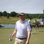 A man in a light blue patterned polo and cap smiles while holding a golf club on a green. Other golfers are in the background, with trees and a pond visible under a clear sky.