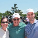Three people smiling outdoors at a golf course, wearing casual clothing and caps, with golf carts and green trees in the background on a sunny day.