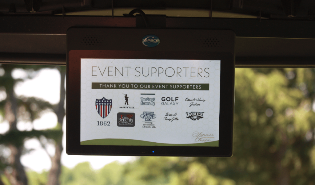 A screen displays a list of event supporters logos and names, including The Great Benefit, Golf Galaxy, Evans Leasing, and several others, with a blurred outdoor background visible behind it.