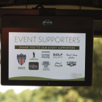 A screen displays a list of event supporters logos and names, including The Great Benefit, Golf Galaxy, Evans Leasing, and several others, with a blurred outdoor background visible behind it.