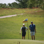 Two people holding golf clubs stand near a flag marked 50 on a golf course, surrounded by green grass and trees in the background under a clear sky.