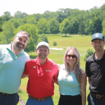 Four adults stand together on a sunny golf course, smiling at the camera. The group includes three men and one woman, all wearing golf attire, with green trees and fairways in the background.