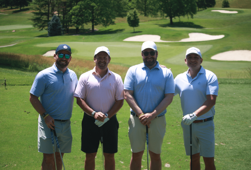 Four men stand side by side on a golf course holding clubs, smiling at the camera. They wear golf attire including caps, polos, and shorts, with sand traps and trees visible in the background.