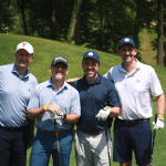 Four men stand together on a sunny golf course, smiling at the camera and holding golf clubs, dressed in polo shirts and caps, with green grass and trees in the background.
