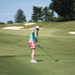 A woman wearing a white cap, turquoise shirt, and red patterned skirt stands on a golf course, holding a golf club and giving a thumbs up toward the camera. Trees and sand traps are visible in the background.