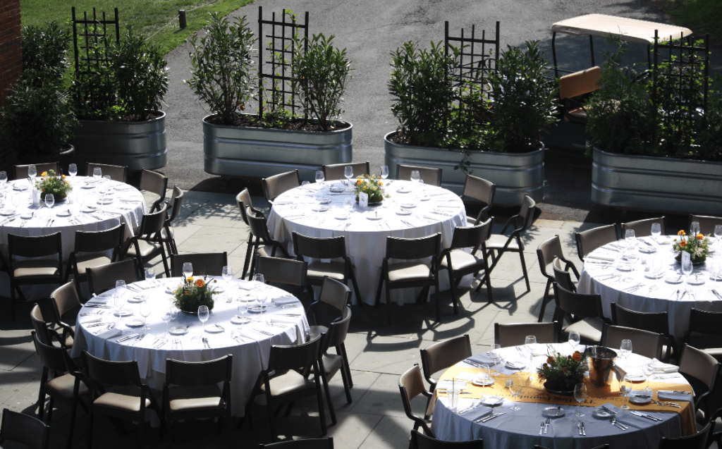 Outdoor event setup with round tables covered in white tablecloths, set with plates and glassware, and decorated with flower centerpieces. Metal planters with green bushes line the background.