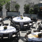Outdoor event setup with round tables covered in white tablecloths, set with plates and glassware, and decorated with flower centerpieces. Metal planters with green bushes line the background.
