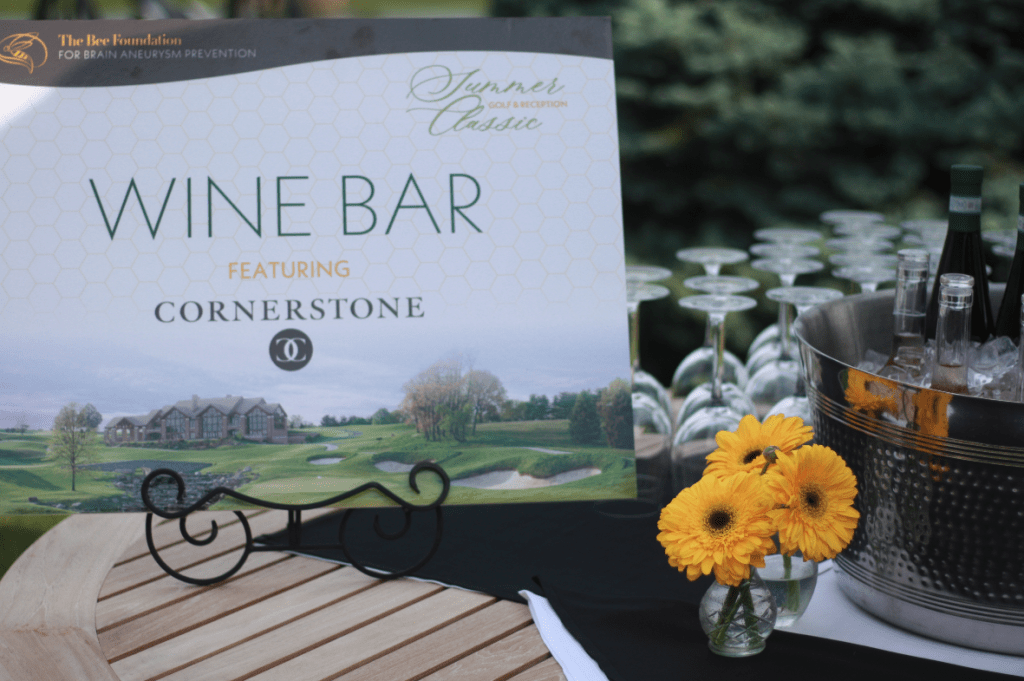 A sign reading Wine Bar featuring Cornerstone is displayed on a table outdoors, alongside a bucket of ice with wine bottles, stacked wine glasses, and a small vase with yellow flowers. Trees are visible in the background.