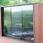 A large window with wooden shutters displays a sign reading Summer Golf & Reception Classic, Tuesday, June 9th, 2016. Green trees and part of a brick building are visible outside.
