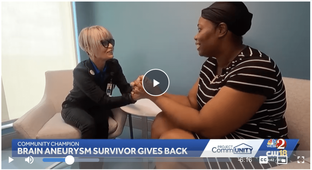 Two women sit facing each other, holding hands and smiling warmly. A text banner reads, COMMUNITY CHAMPION: BRAIN ANEURYSM SURVIVOR GIVES BACK with news logos in the bottom right corner.