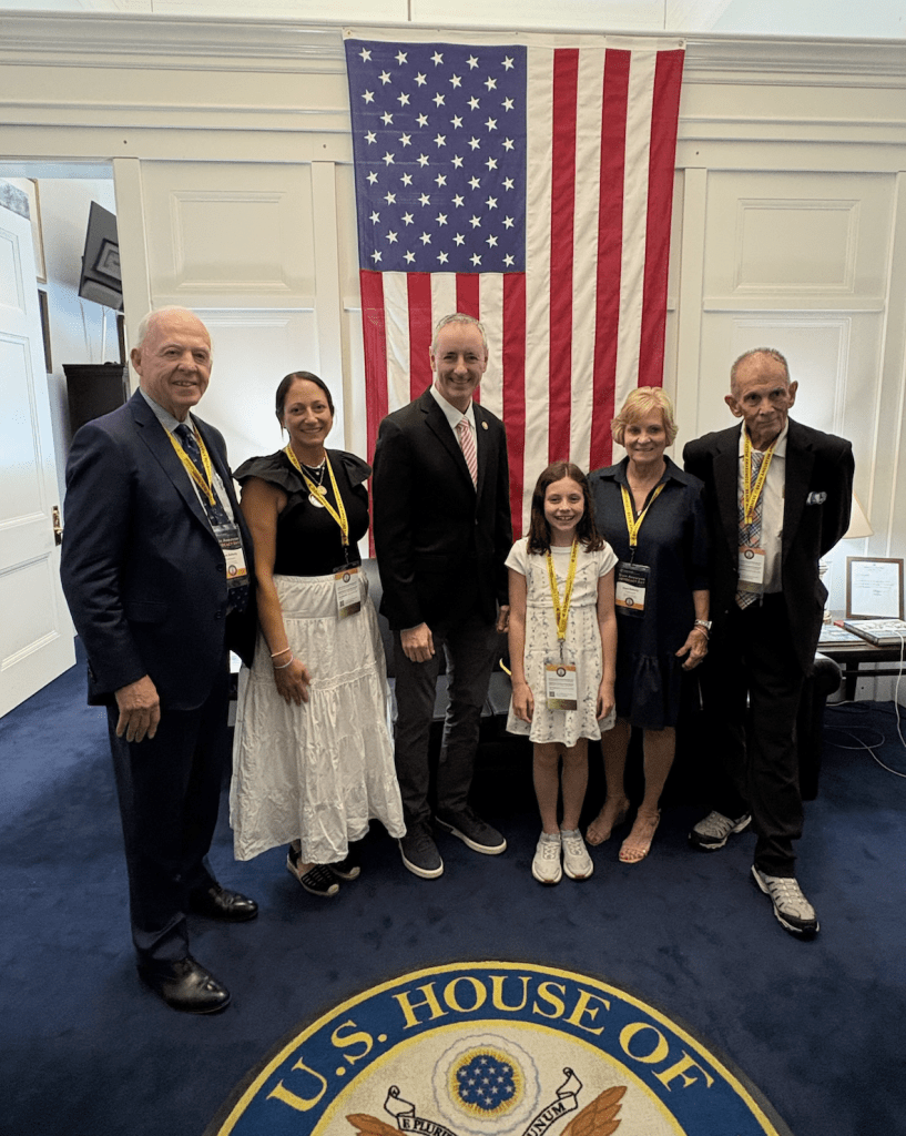 Six people stand in an office with a large U.S. flag hanging behind them and a U.S. House of Representatives seal on the floor. They are smiling and wearing name badges, posing for a group photo.