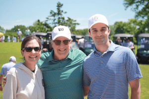 Three people smile outdoors on a sunny day, standing close together. Two men wear baseball caps and polo shirts; the woman wears sunglasses and a light hoodie. Golf carts and trees are visible in the background.
