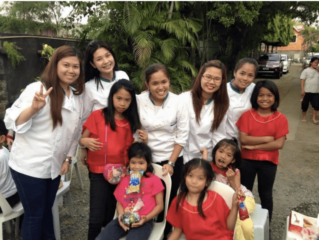 A group of women and young girls, some in white shirts and others in red, smile and pose outdoors. Several girls hold snacks and gifts while sitting on white chairs. Trees and parked cars are visible in the background.