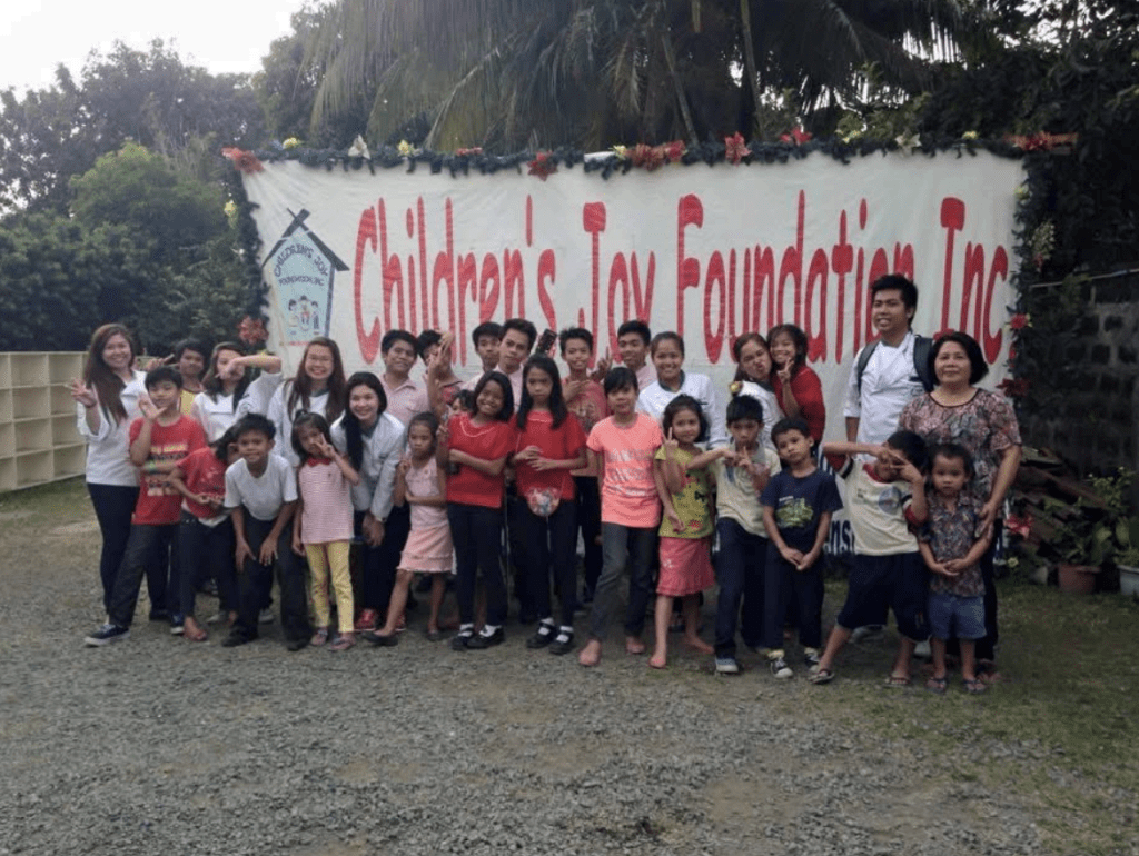 A group of children and adults smile and pose in front of a sign reading Childrens Joy Foundation Inc. outdoors, with trees and plants in the background.