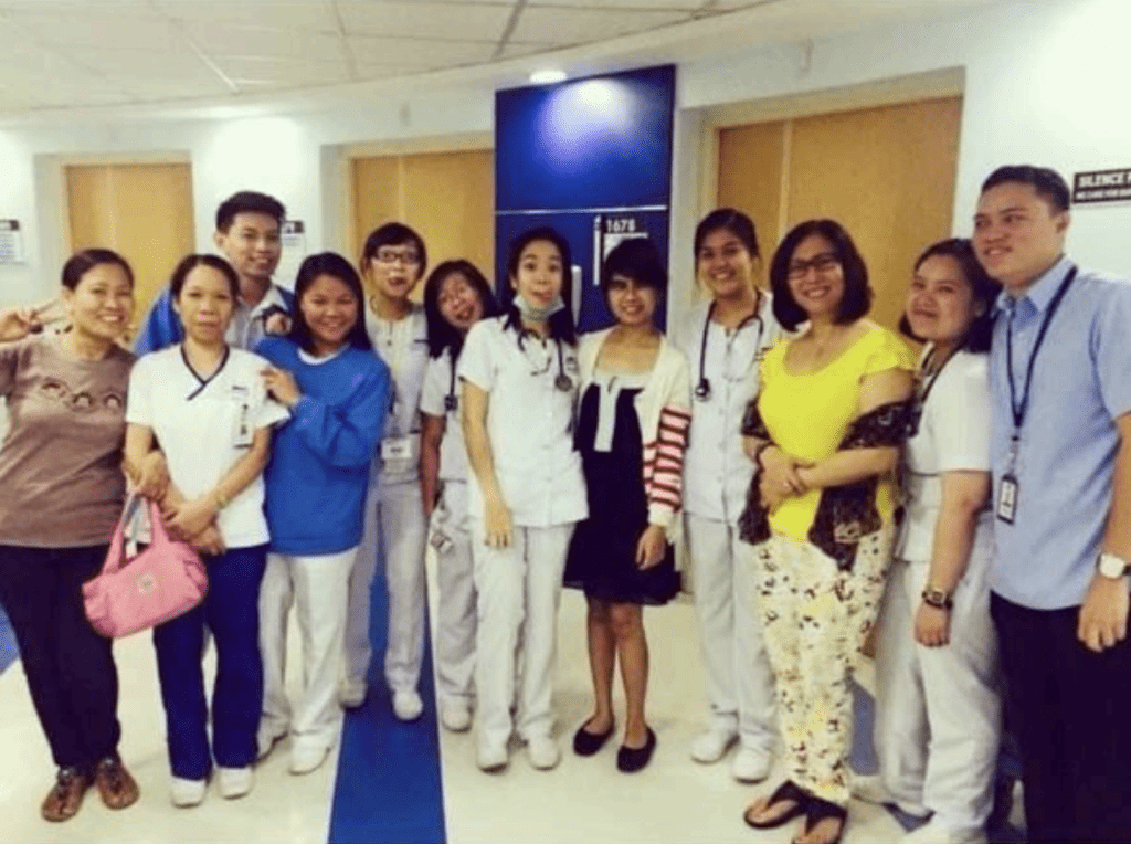 A group of people, some in white medical uniforms and others in casual clothes, smile and pose together in a hospital hallway.