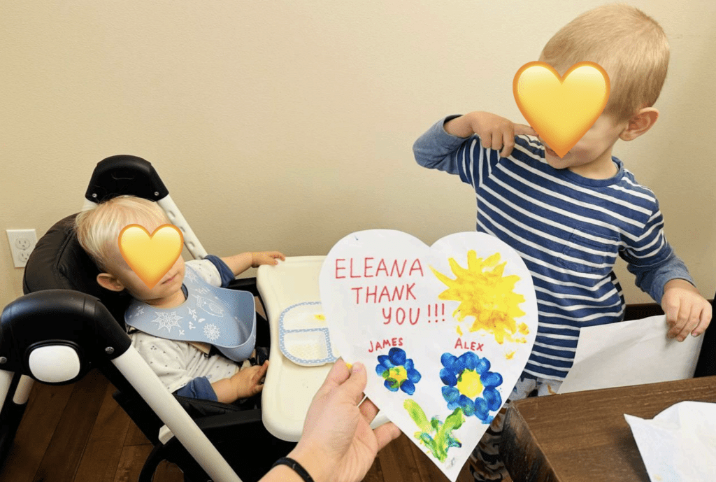 Two young children sit at a table. One is in a high chair, the other stands nearby. A hand holds a heart-shaped card that reads ELEANA THANK YOU!!! with colorful flowers and the names James and Alex.