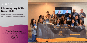 A group of people pose indoors holding booklets, standing behind a banner for The Bee Foundation for Brain Aneurysm Prevention. A screen shows others joining virtually. Text reads “Choosing Joy With Susan Hall” and thanks her for speaking.
