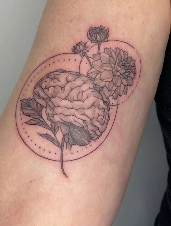 A black and grey tattoo on an arm shows a human brain merging with blooming flowers, surrounded by dotted and solid circles, blending anatomical and botanical elements.