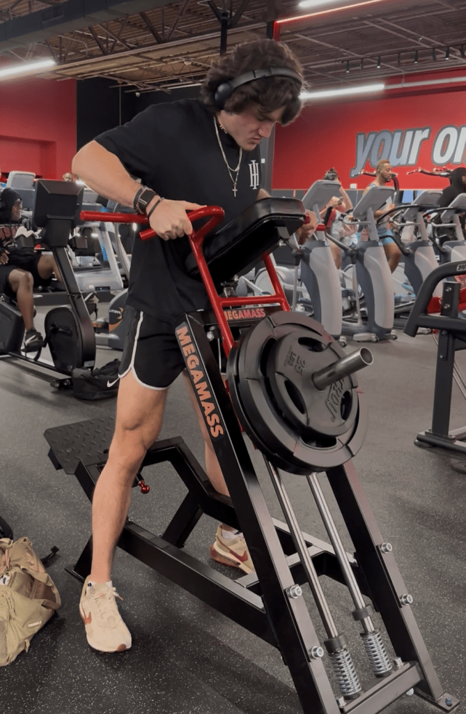 A person in a gym uses a Viking Press machine loaded with weights. Cardio equipment and other people are visible in the background. The person wears a black shirt, black shorts, headphones, and beige sneakers.