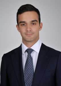 A man in a dark suit, white shirt, and patterned tie poses for a professional headshot in front of a plain light gray background.