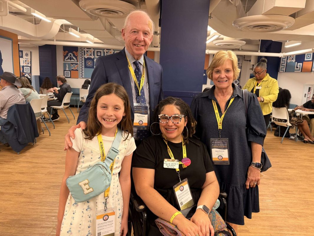 Four people pose and smile together at an indoor event. Two adults stand behind a young girl and a woman in a wheelchair. All are wearing event badges and lanyards. Other attendees are visible in the background.
