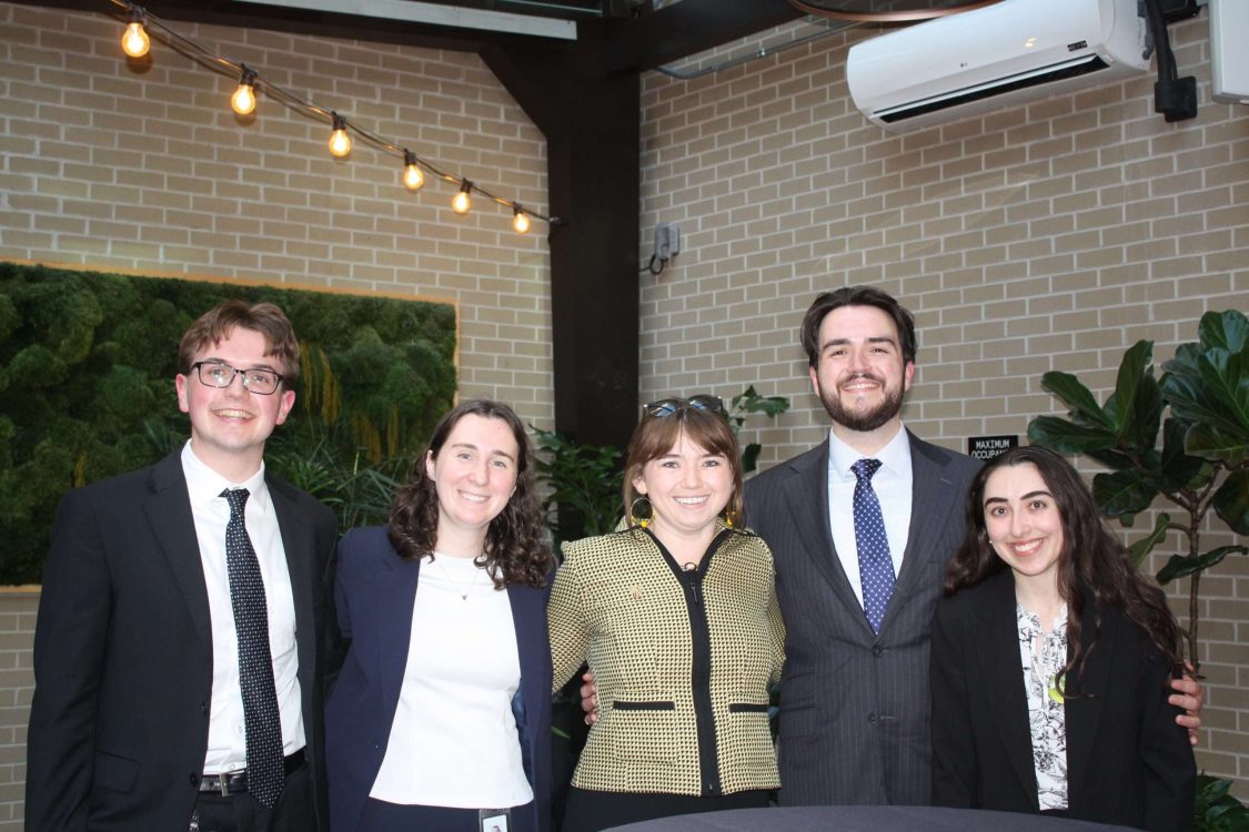 Five people dressed in business attire stand together smiling in front of a brick wall with plants and string lights overhead, posing for a group photo at what appears to be a professional event.