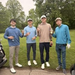 Four young men in golf attire stand on a path by a grassy area, smiling and holding drinks. Trees and green grass are in the background, and it appears to be an overcast day.