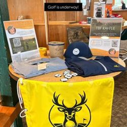 A round table displays golf-themed raffle items, including a yellow flag with a deer logo, hats, shirts, flyers, and a small raffle ticket bucket. A sign in the background reads, Golf is underway!.