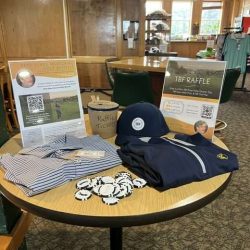 A round table displays raffle prizes including a striped shirt, a navy TBF hat, a navy pullover, golf ball markers, and raffle tickets. Two informational signs about a golf tournament and raffle stand behind the items.