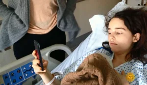 A young woman in a hospital bed, wearing a hospital gown and holding a phone, looks at the screen. Another person stands nearby, partially out of frame. The woman appears calm and is covered with a brown blanket.
