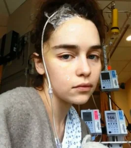 A young woman in a hospital gown sits in a medical setting with IV lines and monitors attached, looking tired. Medical equipment is visible in the background.