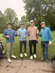 Four young men in golf attire stand on a path by a grassy area, smiling and holding drinks. Trees and green grass are in the background, and it appears to be an overcast day.