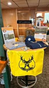 A round table displays golf-themed raffle items, including a yellow flag with a deer logo, hats, shirts, flyers, and a small raffle ticket bucket. A sign in the background reads, Golf is underway!.