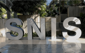 Large, 3D block letters spelling SNIS stand on a polished floor in an indoor atrium. The letters are grey and white with shadows, set against a background of lush potted palm trees and light-colored wooden wall panels.