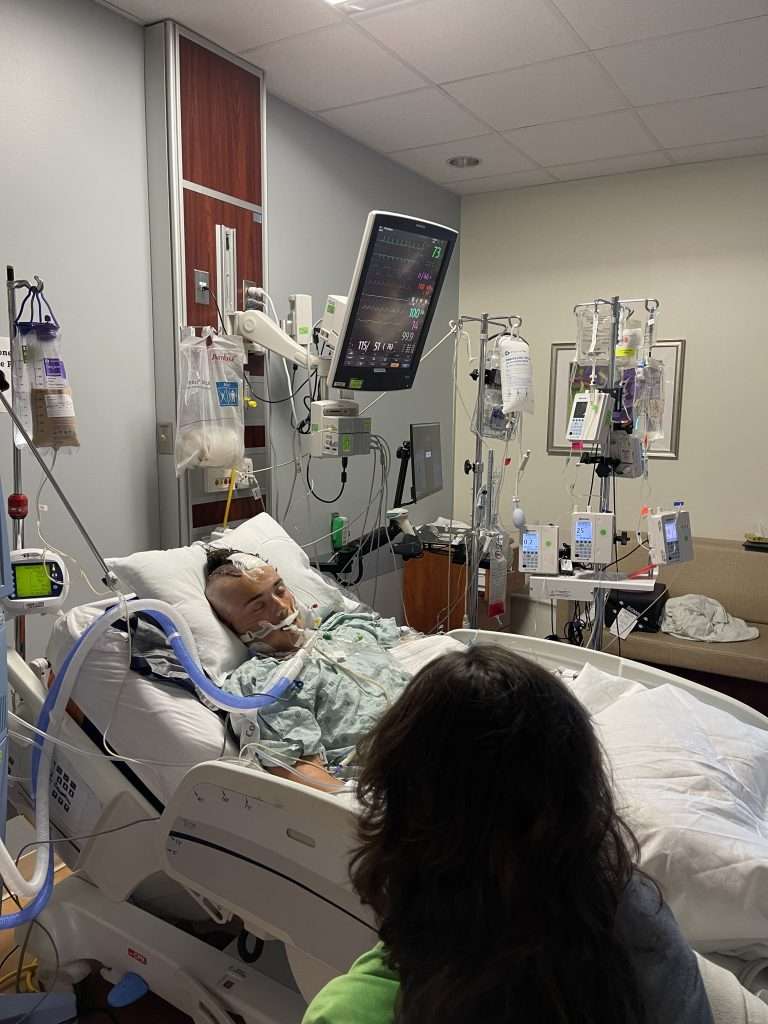 A patient lies in a hospital bed connected to medical devices and monitors, with a visitor sitting nearby. The room contains various IV drips, machines, and a large monitor displaying vital signs.